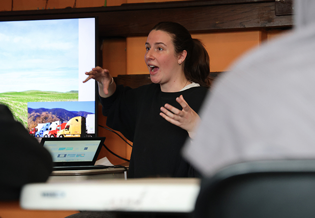 A young woman animatedly teaches in a classroom in front of a screen