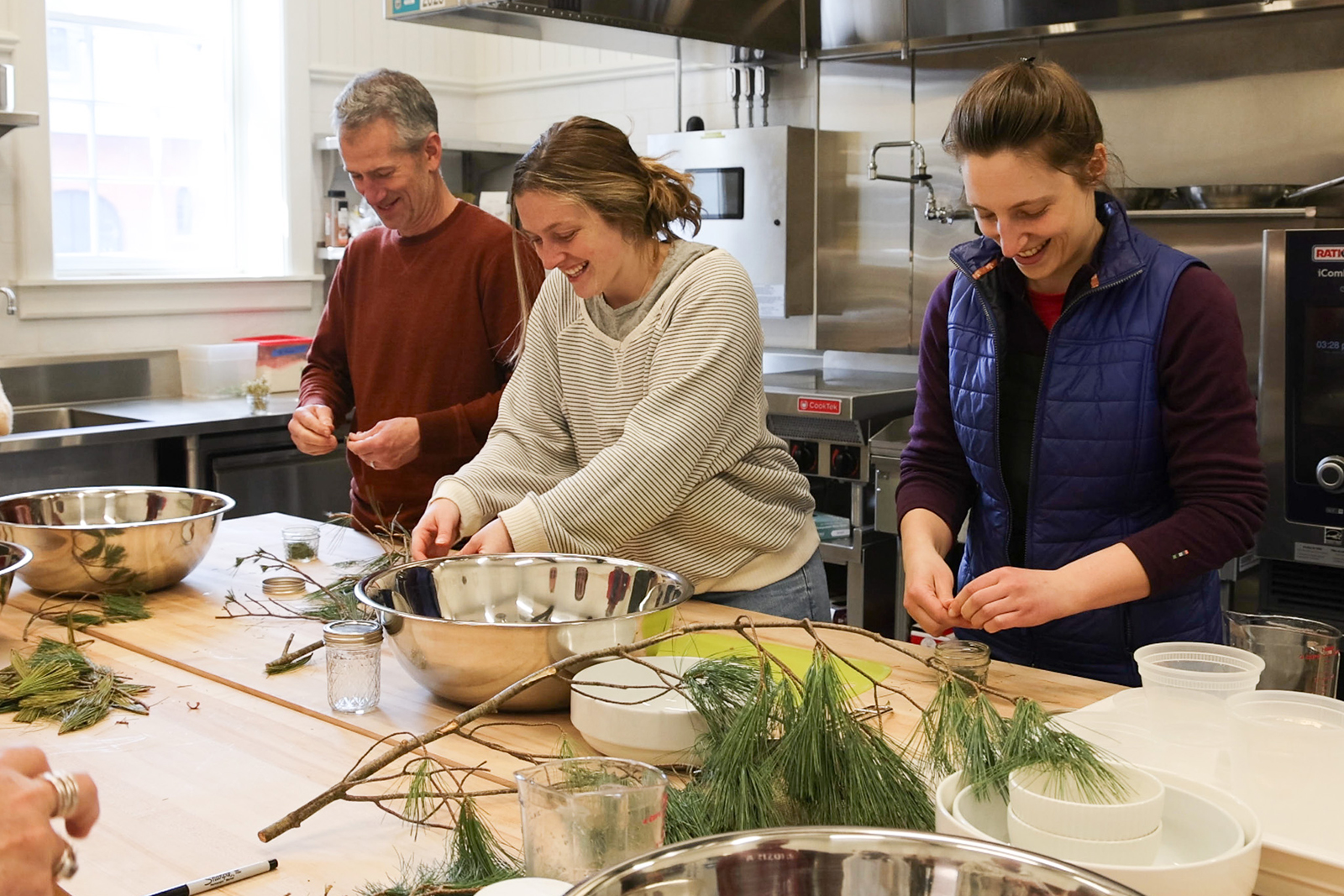 Three adults smile while preparing jars of pine infused honey in an industrial kitchen