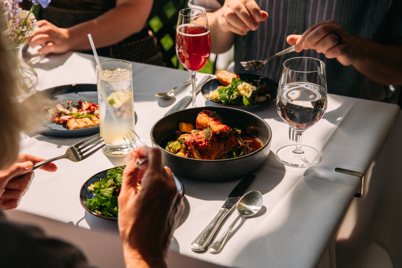 A tight shot of a table shows the hands of two diners holding utensils. Between them are plates of salad, cocktails, and a chicken entrée.