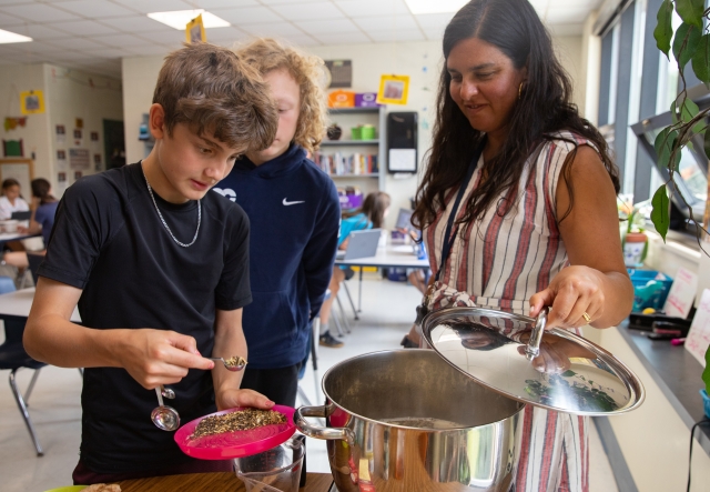 A teacher holds the lid open of a simmer pot of chai as a student spoons in loose herbs and spices