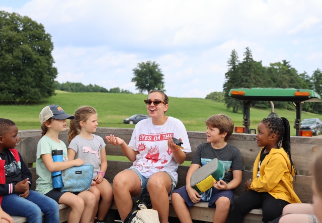A woman rides a tractor pulled wagon with a handful of young summer campers