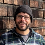 Bearded man with dark hair in a beanie, glasses and a plaid shirt smiles in front of a shingled wall.